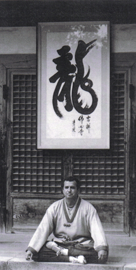 Master Ken Corona seated beneath calligraphy in traditional dobok